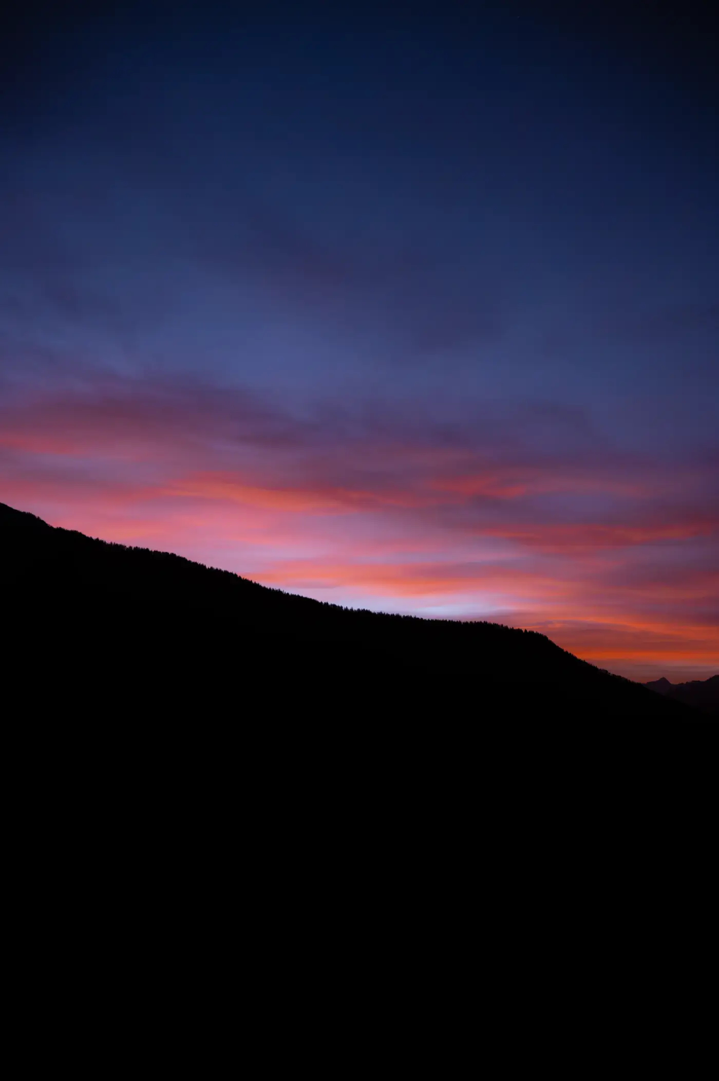 Crête boisée en silhouette noire sous un ciel crépusculaire dégradé de bleu, rose et orange